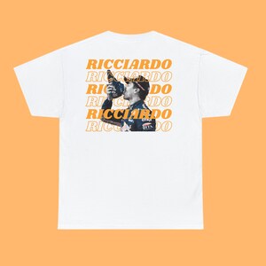 May include: White t-shirt with a repeated pattern of the word "RICCIARDO" in orange text. A photo of a race car driver is centered on the shirt.