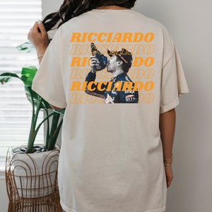 May include: A beige t-shirt with a repeated pattern of the word "Ricciardo" in orange. A photo of a race car driver drinking from a bottle is centred on the shirt.