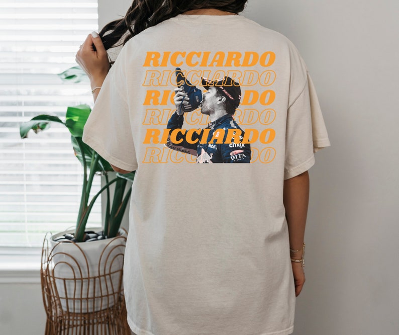 May include: A beige t-shirt with a repeated pattern of the word "Ricciardo" in orange. A photo of a race car driver drinking from a bottle is centred on the shirt.