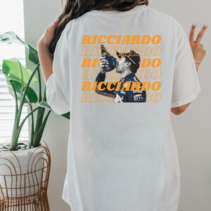 May include: White t-shirt with a repeated image of a race car driver celebrating with a bottle of champagne. The text "RICCIARDO" is repeated in orange on the shirt.