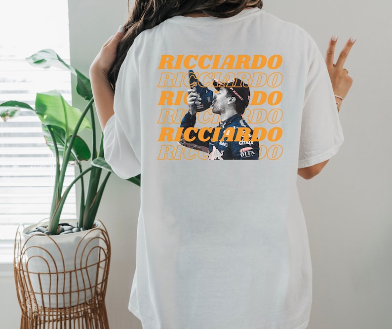 May include: White t-shirt with a repeated image of a race car driver celebrating with a bottle of champagne. The text "RICCIARDO" is repeated in orange on the shirt.