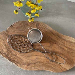 Puede incluir: Dos utensilios de cocina plateados sobre una tabla de cortar de madera. Los utensilios son un colador de malla de alambre y un pequeño colador redondo con mango.