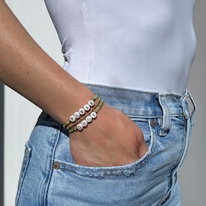 Könnte beinhalten: Eine Person trägt ein weißes Tanktop und eine blaue Jeans mit zwei Perlenarmbändern am Handgelenk. Die Armbänder buchstabieren die Wörter "PIXIE DUST" in weißen Perlen auf einem grünen und goldenen Perlenhintergrund.