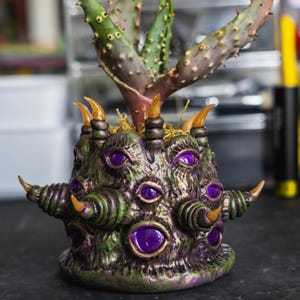 May include: A textured, dark green and purple planter featuring multiple embedded purple eyes and protruding horn-like spikes. A spiky, green succulent plant with reddish-brown tips grows from the center of the detailed, creature-like vessel.