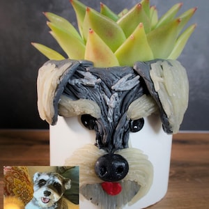 May include: A white cylindrical planter featuring a sculpted, textured gray and cream dog face with black eyes, a black nose, and a red tongue. A green succulent plant grows from the top. A small reference photo of a dog is attached to the side.