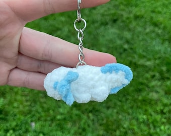 May include: A handmade cloud-shaped keychain. The keychain features a fluffy white body with light blue accents. The keychain is attached to a silver metal ring and chain. The background is blurred green grass.