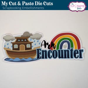 May include: Die-cut embellishment featuring a cartoon depiction of Noah's Ark, a rainbow, and the words "Ark Encounter." The ark is brown with blue water, rain, and gray clouds. The rainbow has red, orange, yellow, green, and blue stripes. Scrapbooking embellishments.