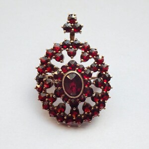 Op de afbeelding: Een vintage goudkleurige broche met een centrale ovale, dieprode edelsteen, omringd door een halo van kleinere, bijpassende rode stenen. De broche heeft een cirkelvormig ontwerp met een decoratief top element.