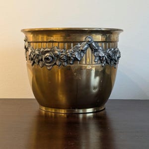Puede incluir: Un macetero de latón dorado con un diseño floral y de uvas plateado alrededor del borde.