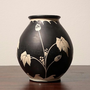 Może przedstawiać: Czarny ceramiczny wazon z kremowym wzorem kwiatowym. Wazon ma zaokrąglony kształt z falistym brzegiem i kremową podstawą. Wzór kwiatowy obejmuje liście, pąki i jagody.