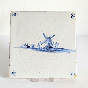 Può includere: Piastrella quadrata in ceramica bianco sporco con una scena di mulino a vento blu. Il mulino a vento è al centro, con un piccolo villaggio sullo sfondo. Accenti floreali blu in ogni angolo. La piastrella è sostenuta da un supporto trasparente.