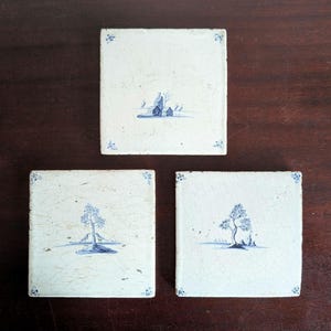 Può includere: Tre piastrelle quadrate in ceramica bianco sporco con disegni di paesaggi blu. Ogni piastrella presenta una scena diversa: una casa, un albero e un albero con una piccola isola. Gli angoli di ogni piastrella hanno piccoli accenti floreali blu.