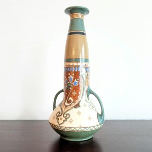 May include: A ceramic vase with two handles, featuring a colourful geometric and floral design in shades of blue, green, brown, and white. The vase has a tall, slender neck and a rounded body.