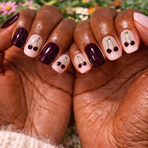 Pode incluir: Close-up de m&atilde;os com unhas cuidadas. Algumas unhas s&atilde;o pintadas de um bord&ocirc; profundo, enquanto outras s&atilde;o rosa claro com um desenho de cereja. O desenho da cereja apresenta duas pequenas cerejas pretas com caules verdes.