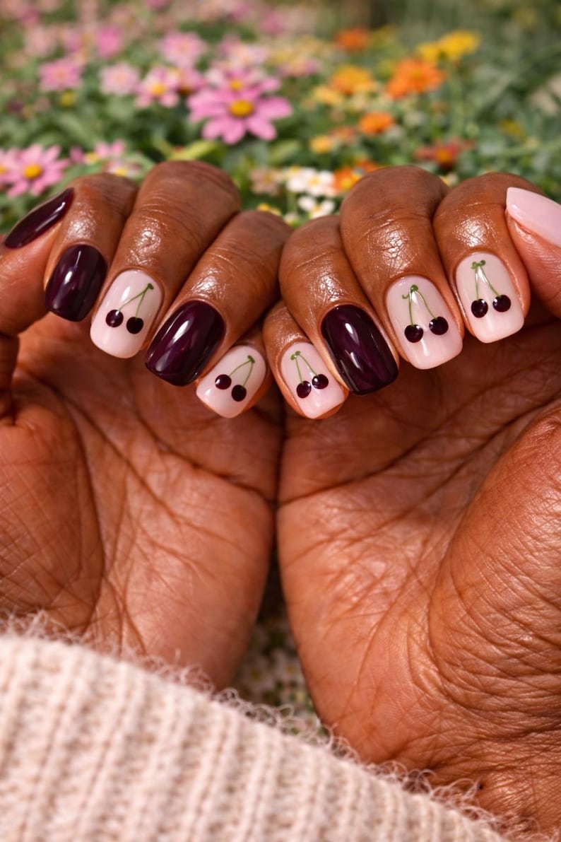 Pode incluir: Close-up de m&atilde;os com unhas cuidadas. Algumas unhas s&atilde;o pintadas de um bord&ocirc; profundo, enquanto outras s&atilde;o rosa claro com um desenho de cereja. O desenho da cereja apresenta duas pequenas cerejas pretas com caules verdes.