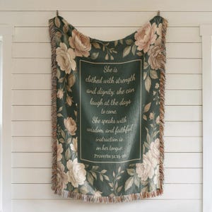Puede incluir: Una manta verde oscuro con un dise&ntilde;o floral y el texto "She is clothed with strength and dignity... Proverbios 31:25-26". La manta tiene un borde de flecos y est&aacute; decorada con rosas y hojas color crema.