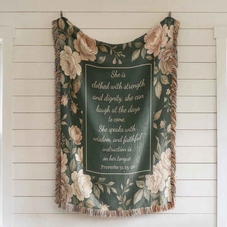 Puede incluir: Una manta verde oscuro con un dise&ntilde;o floral y el texto "She is clothed with strength and dignity... Proverbios 31:25-26". La manta tiene un borde de flecos y est&aacute; decorada con rosas y hojas color crema.