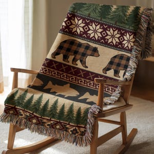 May include: A woven throw blanket with a rustic design featuring bears, trees, and geometric patterns. The blanket is draped over a wooden rocking chair. The color palette includes beige, brown, green, and burgundy.