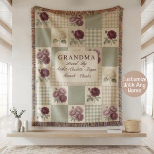 May include: A personalized throw blanket with a patchwork design featuring floral patterns and gingham squares in shades of cream, sage green, and burgundy. The text "GRANDMA Loved By" is displayed, along with names. The blanket has a fringed edge.