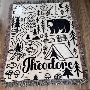 May include: A cream-colored woven throw blanket featuring a black line-art design of a camping scene with a bear, a tent, a campfire, pine trees, fish, and the name Theodore written in a large script font, finished with a fringed edge.