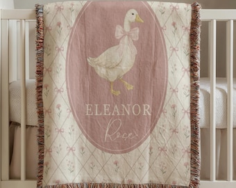May include: A woven throw blanket featuring a central oval frame with a white goose wearing a pink bow. The name Eleanor Rose is printed below the goose. The background has a cream-colored diamond lattice pattern with small pink flowers.