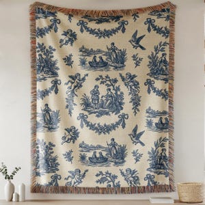 Puede incluir: Una manta crema con un estampado azul toile de Jouy con querubines, figuras, p&aacute;jaros y guirnaldas florales. La manta tiene un borde con flecos en tonos marrones y naranjas. El dise&ntilde;o general es de estilo cl&aacute;sico y decorativo.