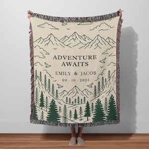 May include: Beige woven throw blanket with a green mountain and forest design. The text "ADVENTURE AWAITS" is above the names "EMILY & JACOB" and the date "09.10.2025". The blanket has a fringed edge.