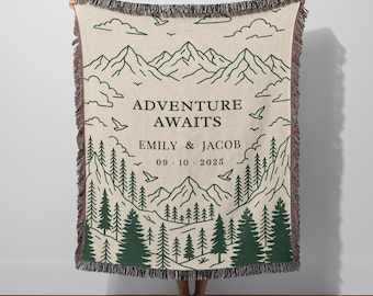 May include: Beige woven throw blanket with a green mountain and forest design. The text "ADVENTURE AWAITS" is above the names "EMILY & JACOB" and the date "09.10.2025". The blanket has a fringed edge.