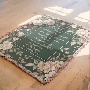 Puede incluir: Una manta tejida con un fondo verde oscuro y un dise&ntilde;o floral con rosas y hojas color crema. El centro de la manta tiene un texto enmarcado en blanco que dice "Ella est&aacute; vestida de fuerza y dignidad... Proverbios 31:25-26."