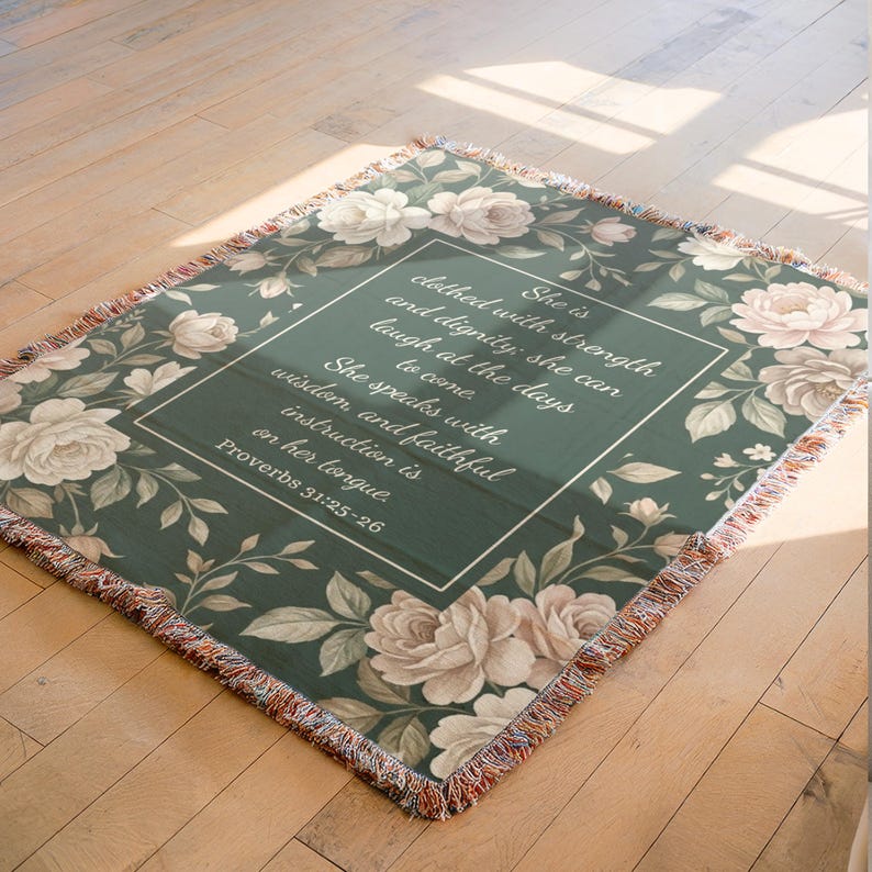 Puede incluir: Una manta tejida con un fondo verde oscuro y un dise&ntilde;o floral con rosas y hojas color crema. El centro de la manta tiene un texto enmarcado en blanco que dice "Ella est&aacute; vestida de fuerza y dignidad... Proverbios 31:25-26."