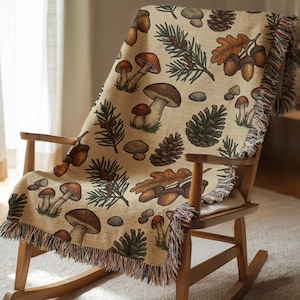 May include: A beige throw blanket with a woodland design, draped over a wooden rocking chair. The blanket displays mushrooms, pinecones, acorns, and leaves in brown, green, and tan. It has a fringed edge.