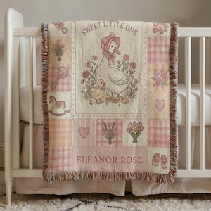 May include: A baby blanket with a patchwork design featuring a goose and duckling illustration, hearts, flowers, and the text "SWEET LITTLE ONE" and "ELEANOR ROSE". The blanket has a pink and cream colour scheme and a fringed edge.
