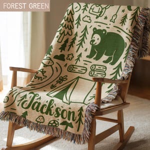 May include: A woven throw blanket in cream with a forest green camping-themed pattern featuring a bear, pine trees, a tent, logs, and fish. The name Jackson is printed in a cursive script at the bottom. The blanket has fringed edges on all sides.