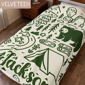 May include: A cream-colored blanket featuring a dark green line-art pattern of a camping scene. The design includes a bear, a tent, pine trees, fish, logs, and a person fishing. The name Jackson is printed in large dark green script letters.