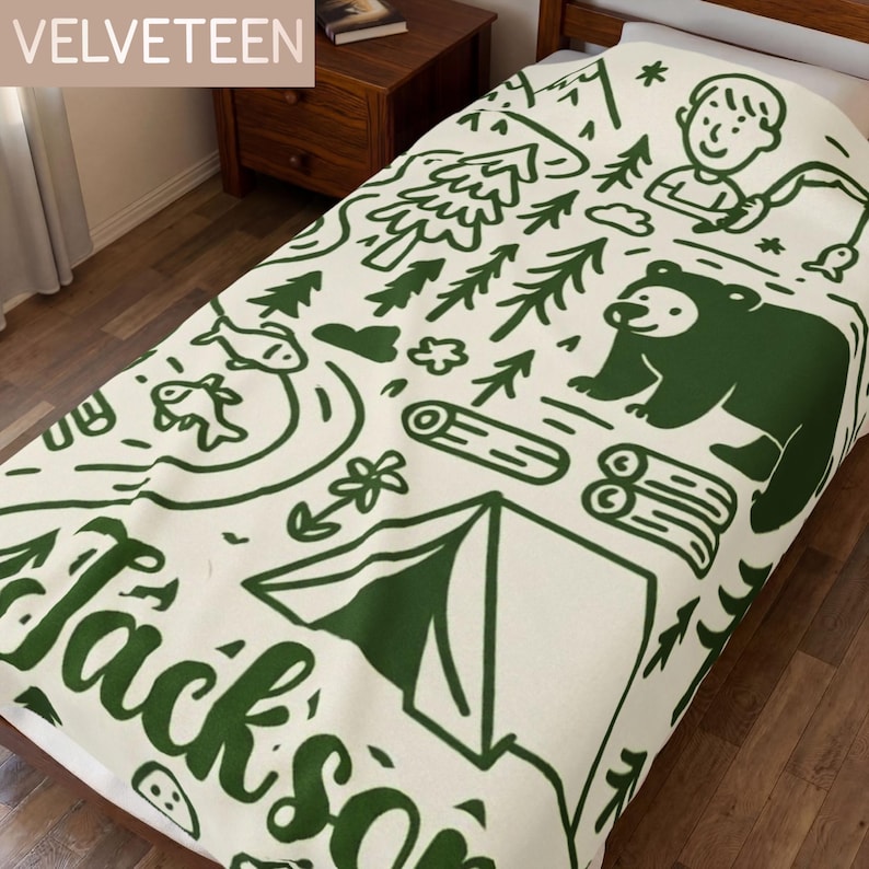 May include: A cream-colored blanket featuring a dark green line-art pattern of a camping scene. The design includes a bear, a tent, pine trees, fish, logs, and a person fishing. The name Jackson is printed in large dark green script letters.