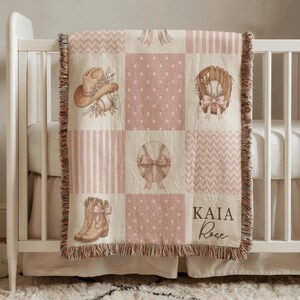 May include: A woven throw blanket featuring a patchwork design in shades of pink and cream. The pattern includes illustrations of a cowboy hat, baseball glove, baseball, and cowboy boots, all accented with bows. The text KAIA Rose is printed.