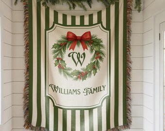 May include: A green and white striped throw blanket with a decorative wreath design and the text "Williams Family." The wreath features a red bow, holly, and berries. The blanket has a fringed edge and is decorated with a garland.