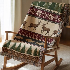 May include: A woven throw blanket draped over a wooden rocking chair. The blanket features a beige background with deer, fish, and tree patterns in brown, green, and burgundy. The blanket has a fringed edge.