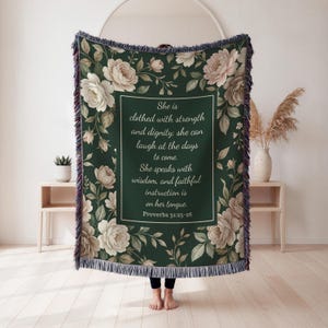 Puede incluir: Una manta verde oscuro con un dise&ntilde;o floral con rosas blancas y color rubor y follaje. La manta tiene un cuadro de texto rectangular con la cita de Proverbios 31:25-26. La manta tiene un borde con flecos.
