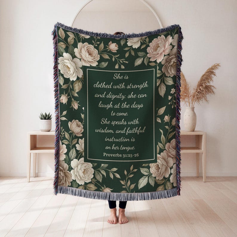 Puede incluir: Una manta verde oscuro con un dise&ntilde;o floral con rosas blancas y color rubor y follaje. La manta tiene un cuadro de texto rectangular con la cita de Proverbios 31:25-26. La manta tiene un borde con flecos.