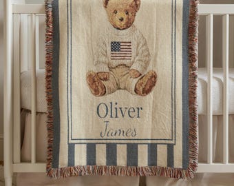 May include: A woven throw blanket featuring a central illustration of a brown teddy bear wearing a cream-colored cable knit sweater with an American flag design. The name Oliver James is printed below the bear. The blanket has dark blue striped borders.