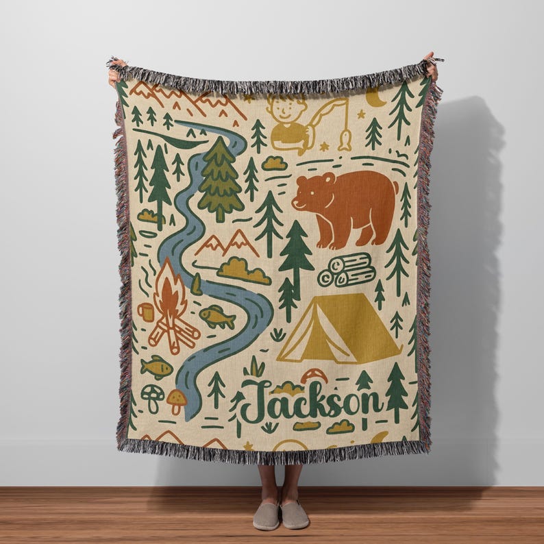 Personalized kids camping blanket with forest illustrations, bear, campfire, river, tent, and custom name Jackson held outdoors in a field.