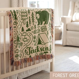 May include: A woven throw blanket in cream with a forest green camping-themed pattern featuring a bear, a tent, trees, a campfire, fish, and mushrooms. The name Jackson is printed in green script. The blanket has a multi-colored fringed edge.