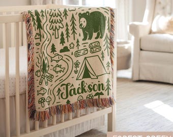May include: A woven throw blanket in cream with a forest green camping-themed pattern featuring a bear, a tent, trees, a campfire, fish, and mushrooms. The name Jackson is printed in green script. The blanket has a multi-colored fringed edge.