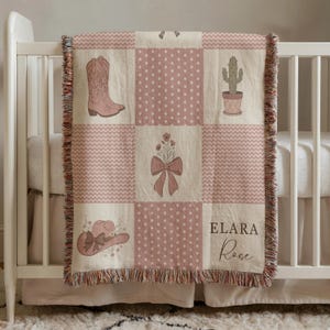 May include: A pink and cream-coloured baby blanket with a patchwork design featuring a cowboy boot, cactus, and a cowboy hat. The blanket has a fringed edge and the name "ELARA Rose" is printed on it.