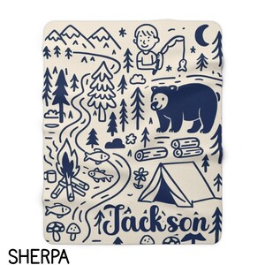May include: A cream-colored sherpa blanket featuring a dark blue line art pattern of a camping scene. The design includes mountains, pine trees, a person fishing, a bear, a campfire, a tent, fish, mushrooms, and the name Jackson in script.
