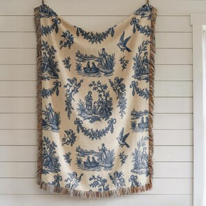 Blue toile woven blanket featuring cherubs, pastoral countryside scenes, and floral garlands in French country farmhouse style.