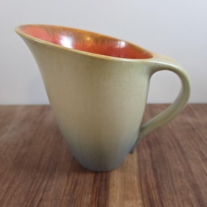May include: A ceramic pitcher with a wide, angled spout and a curved handle. The exterior features a smooth gradient transition from a pale beige at the top to a muted grey at the base. The interior is finished in a vibrant, glossy orange glaze.