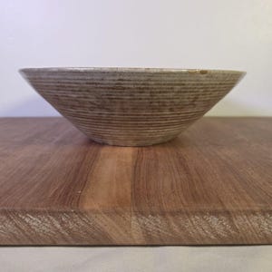 May include: A wide, shallow ceramic bowl featuring a textured, ribbed exterior with a speckled beige and brown glaze. The bowl has a tapered, conical shape with a smooth, slightly lighter-colored interior rim and a visible chip on the edge.