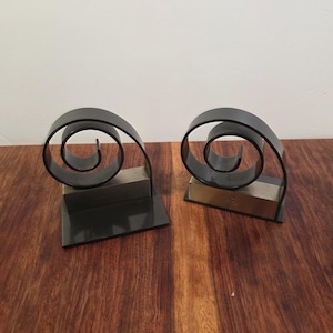 May include: A pair of bookends featuring a black metal spiral design mounted on a rectangular base. Each piece includes a metallic silver-toned block section at the front, providing a contrast to the dark, curved, coiled structure of the metal.